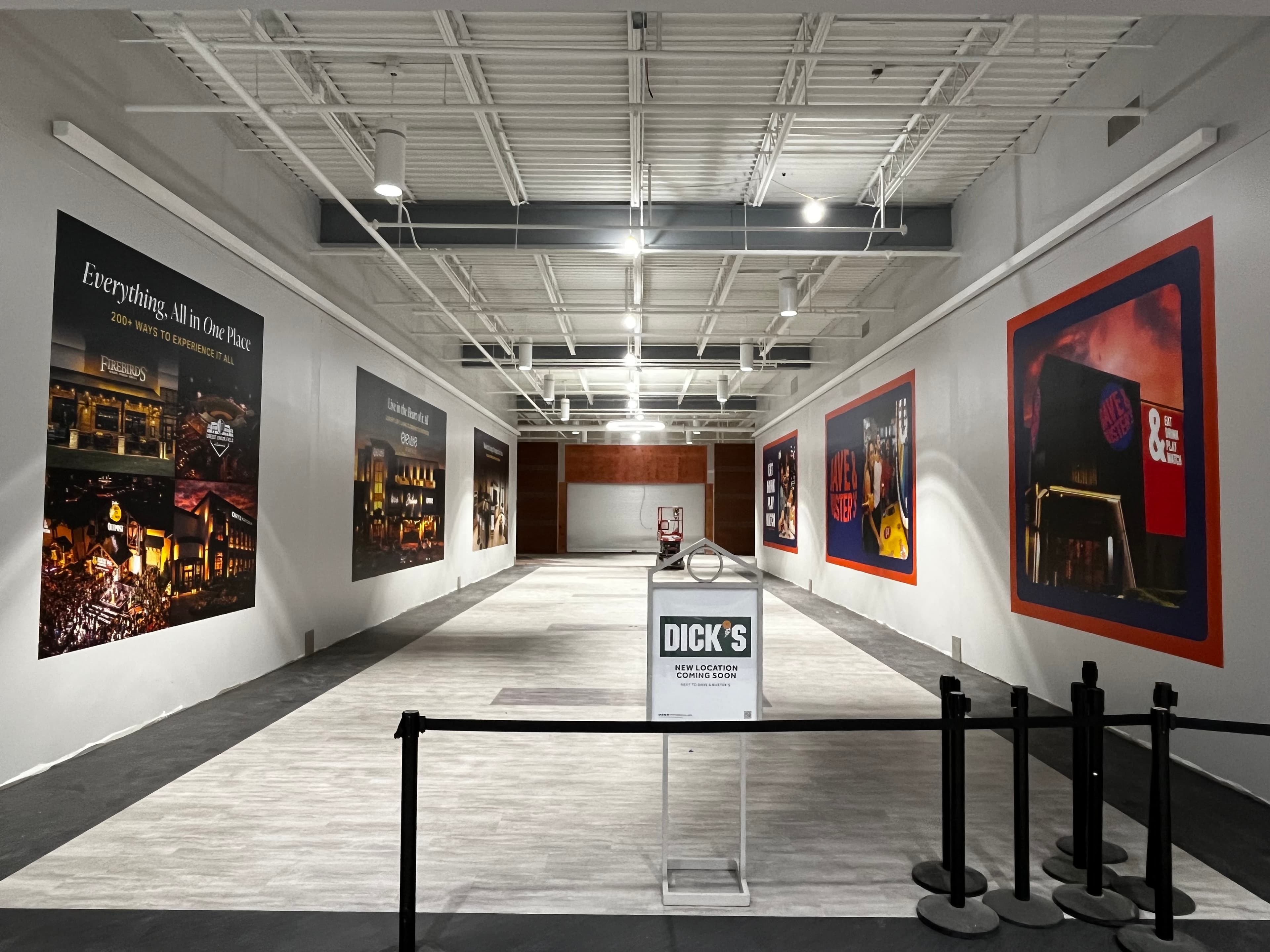 Hallway lined with multi-panel tenant wall graphics and a coming-soon stanchion in the foreground